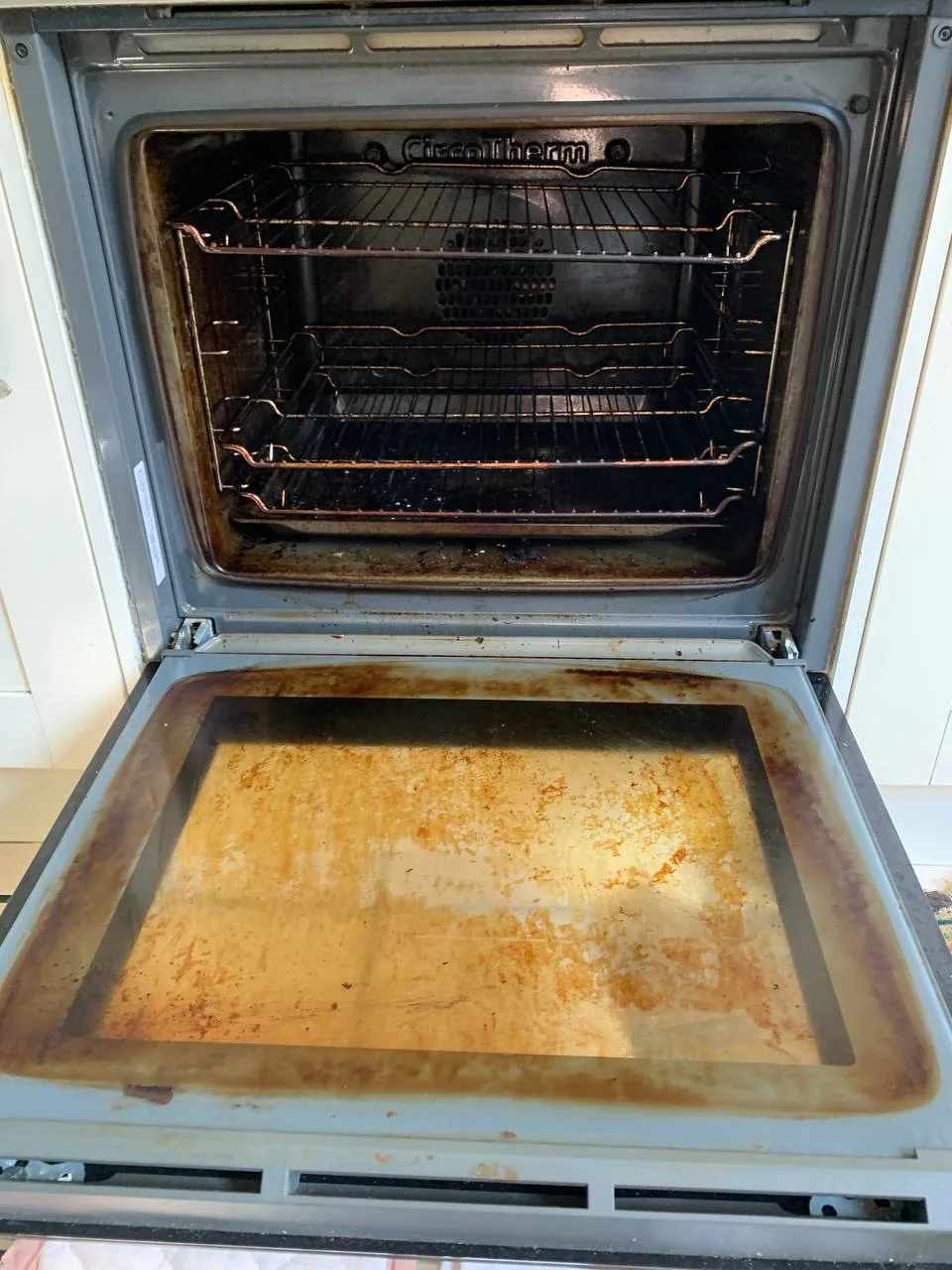 Oven with the door open, thick brown baked-on grease on the inner door glass and dark racks.
